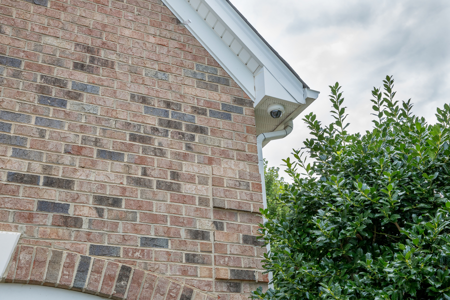 Outdoor security camera mounted under the eaves of a brick home, partially hidden by greenery and pointed downward.